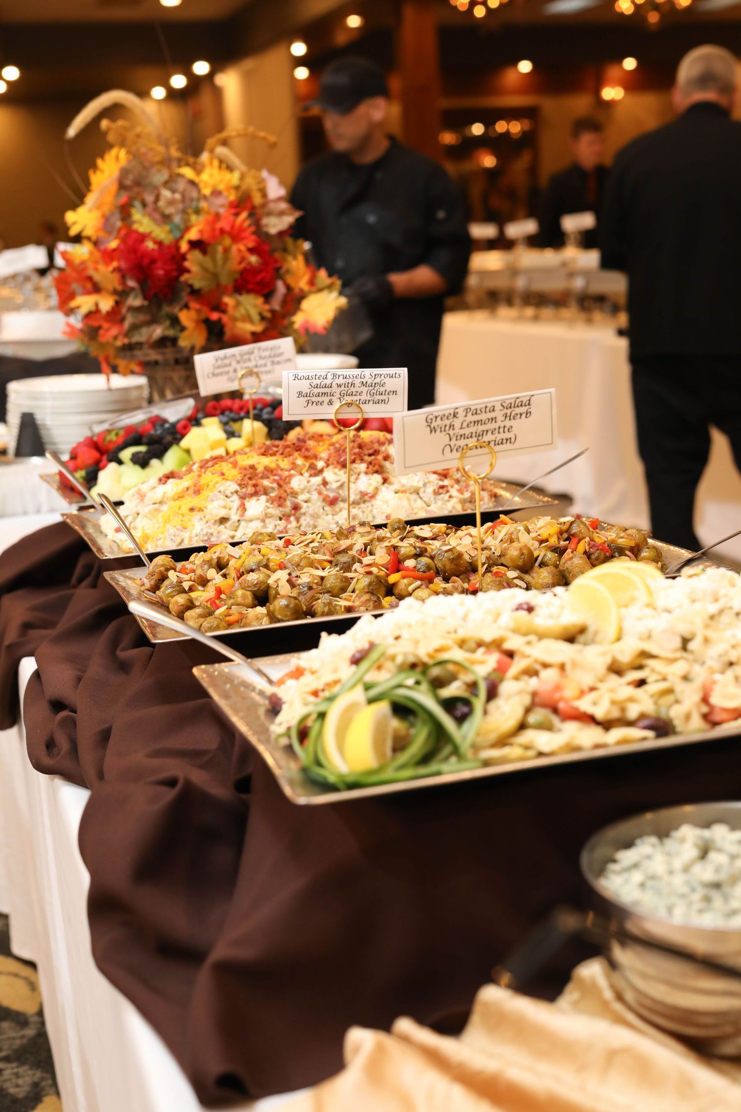 Elegant buffet table with Greek pasta salad, stuffed olives, and fresh vegetable platters beside a fall floral centerpiece as chefs look on.