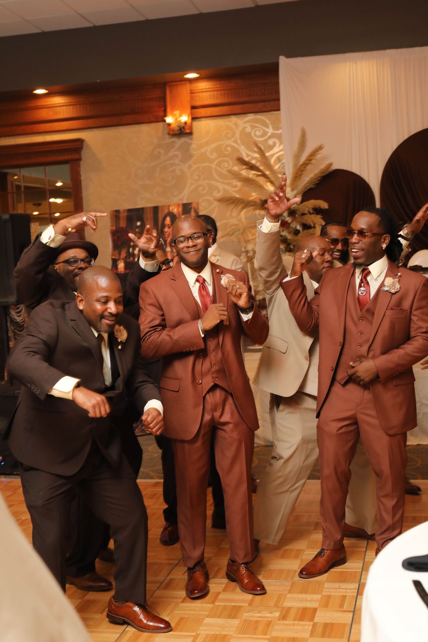 Group of men in brown and tan suits dancing joyfully and pointing at a festive indoor wedding reception.