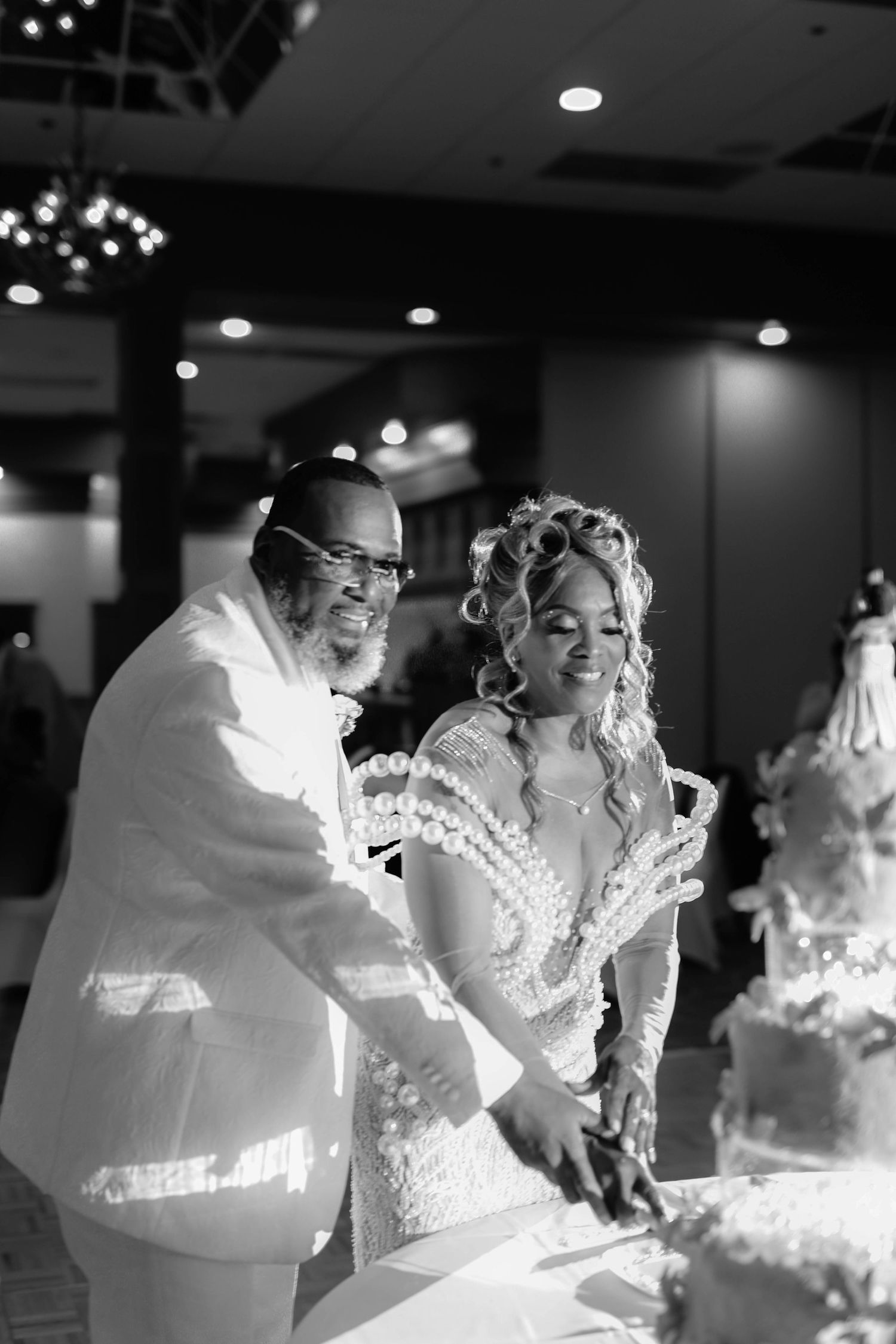Black and white photo of a bride and groom smiling as they cut their tiered wedding cake together at an elegant reception.