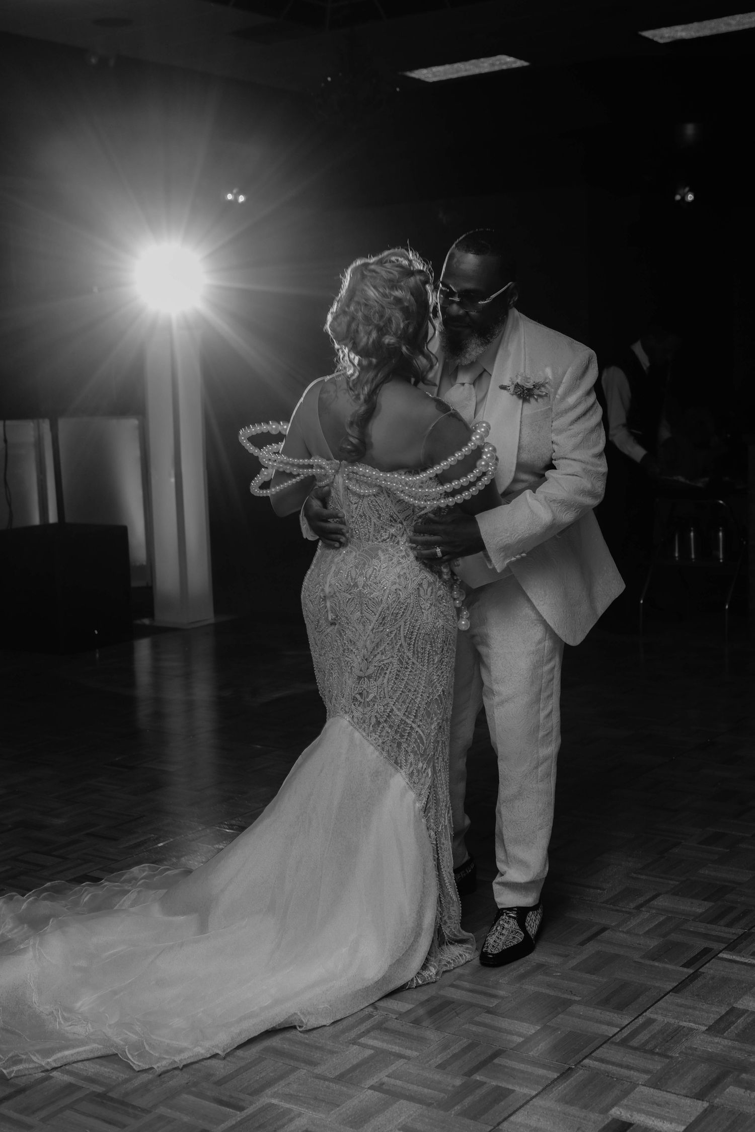 Black and white photo of a bride in a beaded mermaid gown with pearl shoulder detail sharing a first dance with her groom in a white suit.
