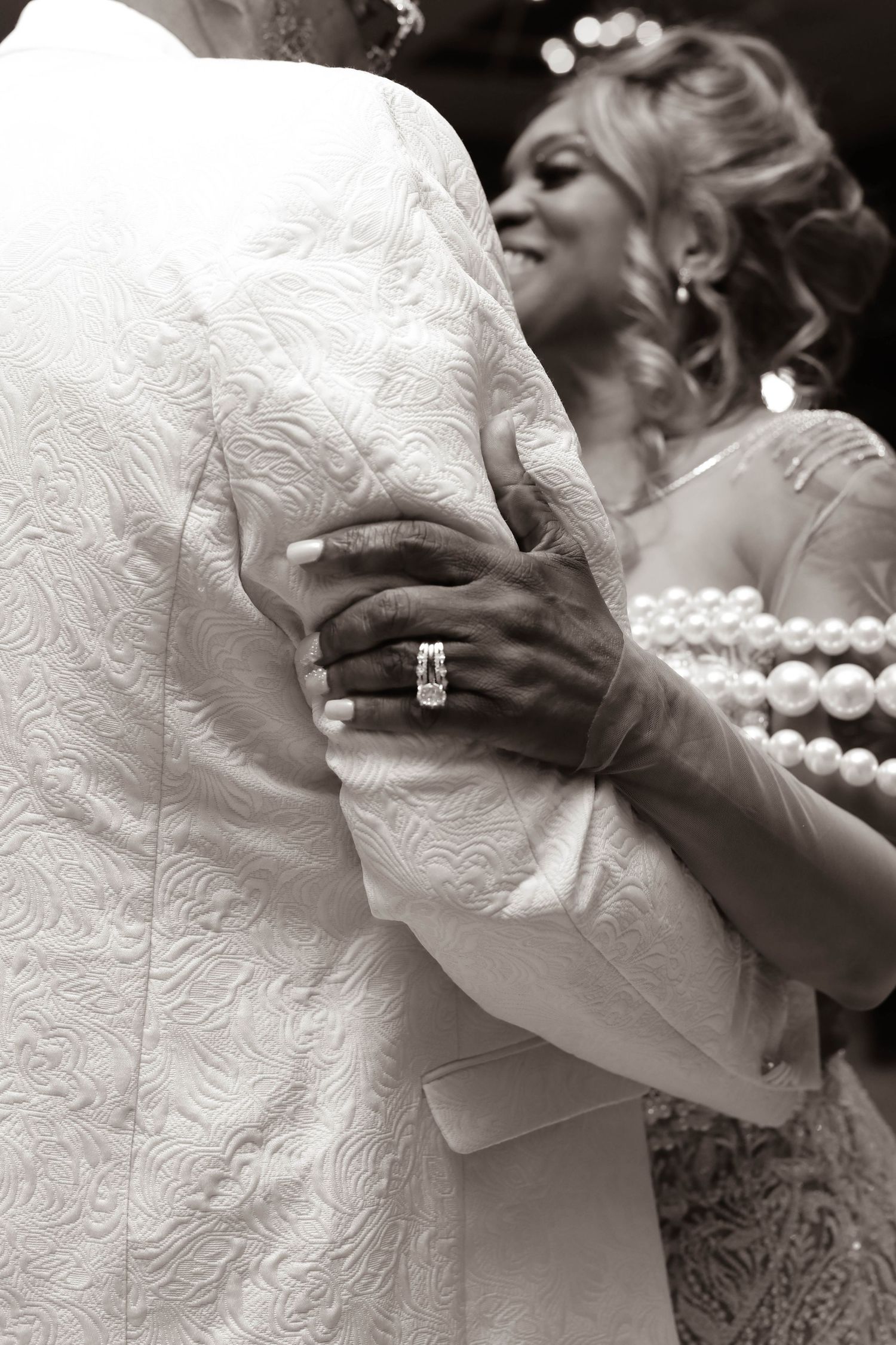 Sepia wedding photo of a couple slow dancing, her hand with a sparkling diamond ring resting on his textured jacket as she smiles.