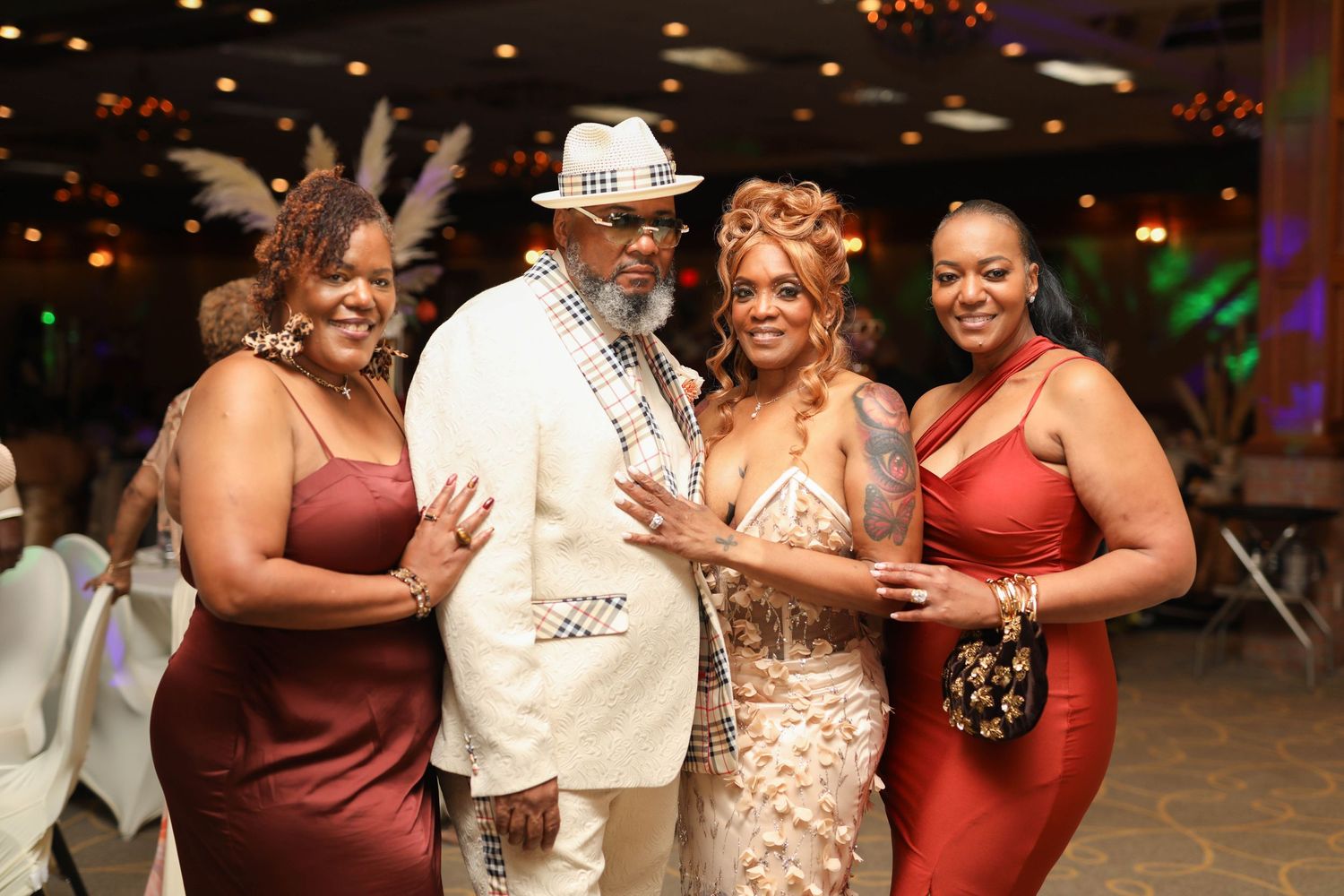 Three smiling women in elegant burgundy, champagne, and red dresses pose with a man in a white suit and hat at a festive banquet event.