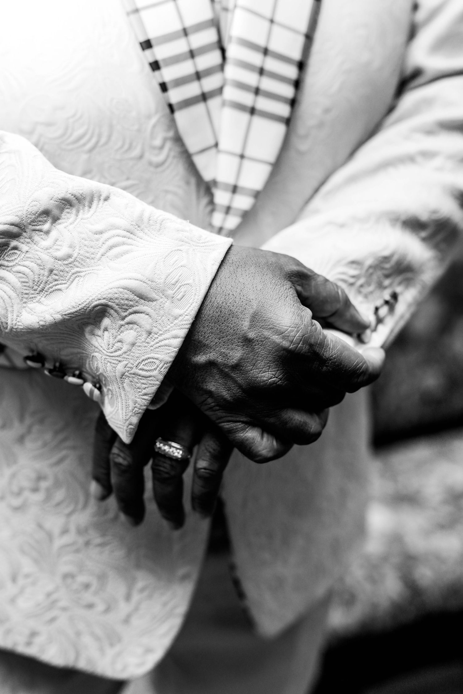 Black and white close-up of a man's clasped hands wearing a ring, dressed in an ornate brocade suit jacket and a plaid tie.