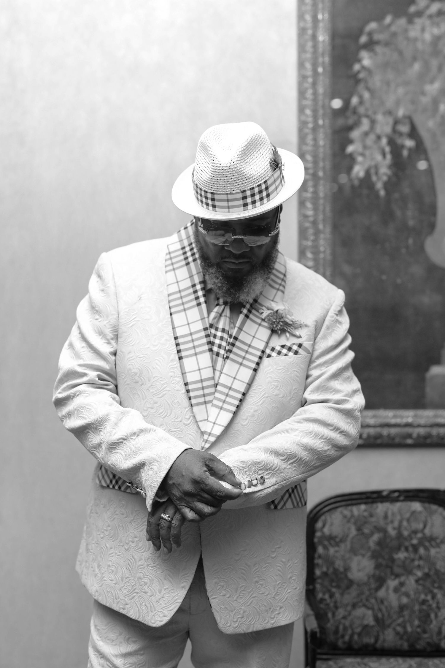 Black and white photo of a bearded man in a textured white blazer with plaid lapels, a plaid fedora and tinted glasses, looking down.