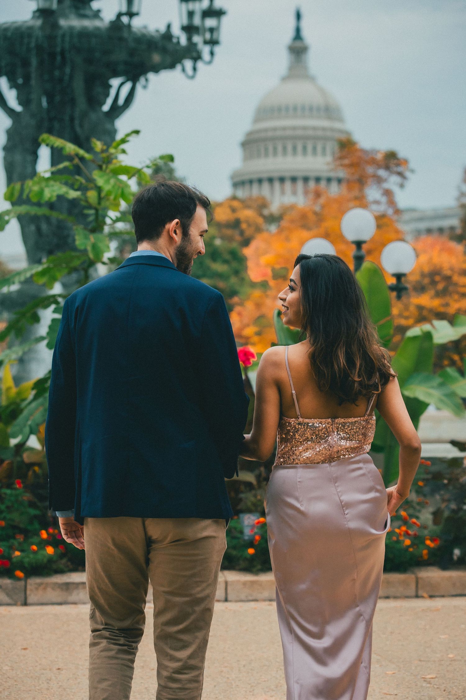 Couple in formal attire walk hand in hand through a garden with colorful autumn foliage, with the U.S. Capitol dome in the background.