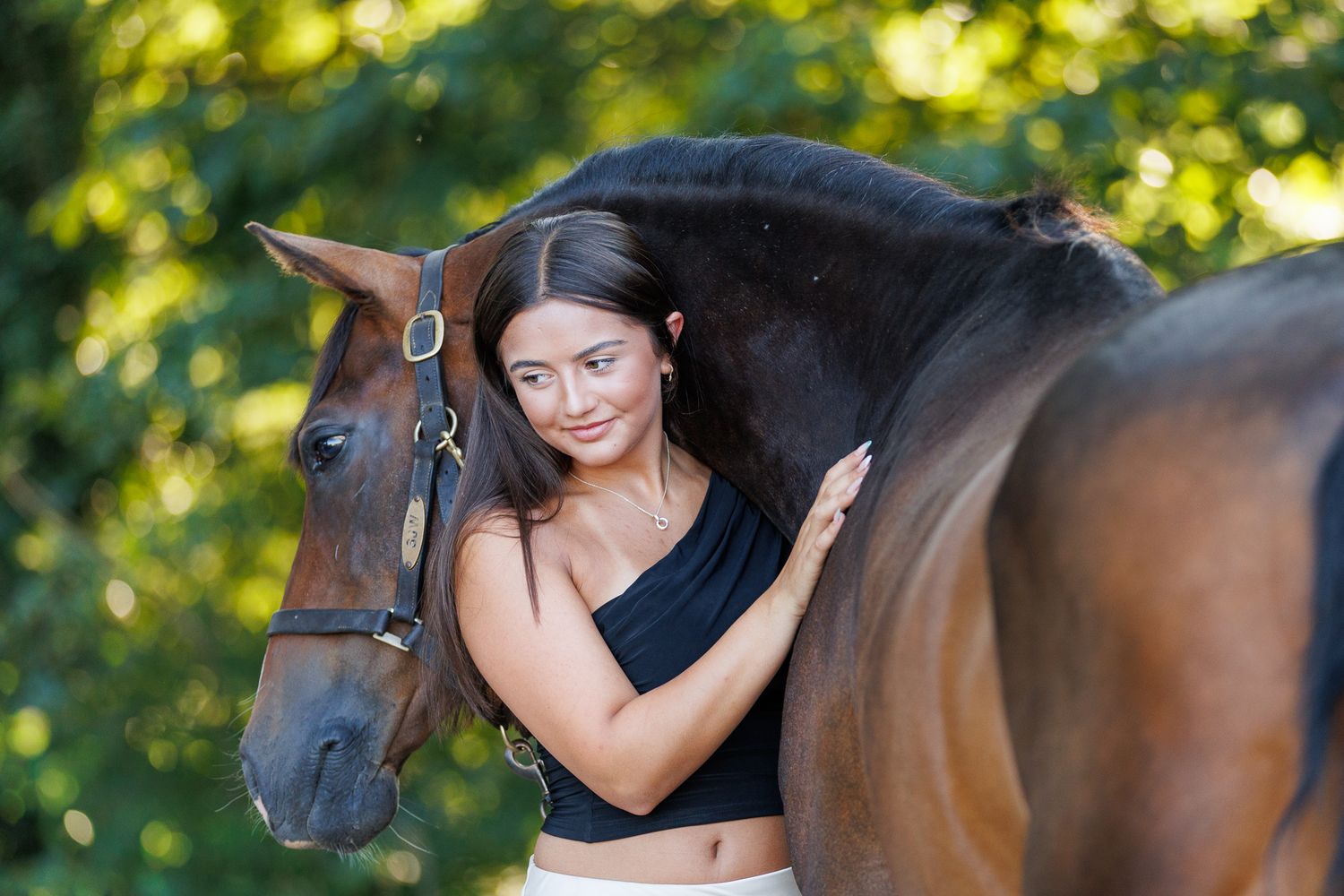 Young woman in black top smiling while gently embracing a brown horse outdoors among lush green trees.
