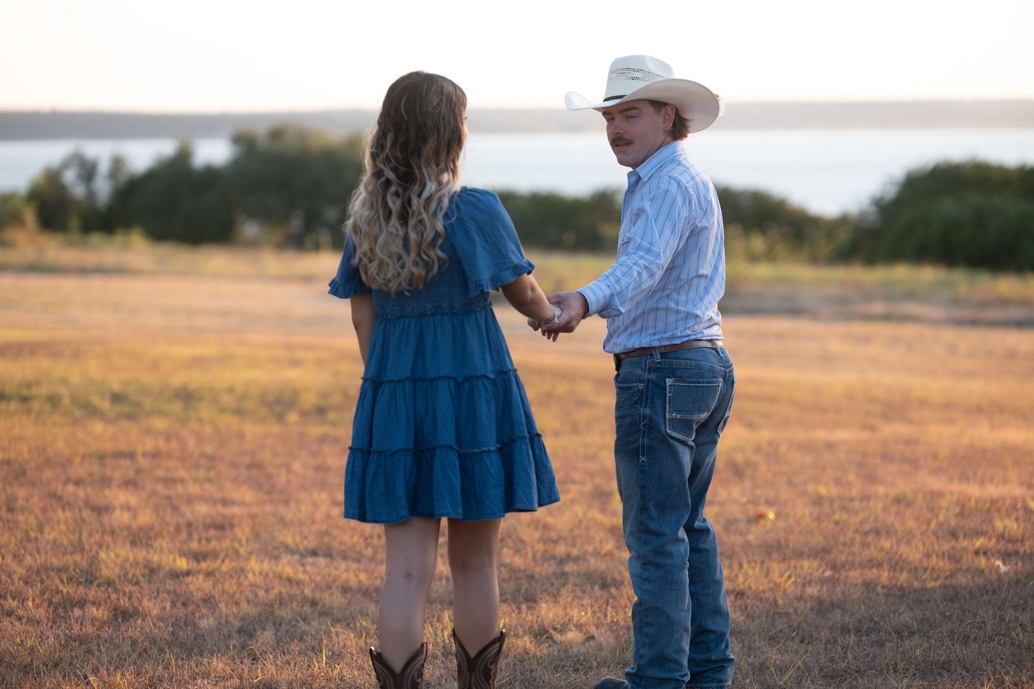 A couple in Western attire holds hands in a golden field near a lake at sunset.