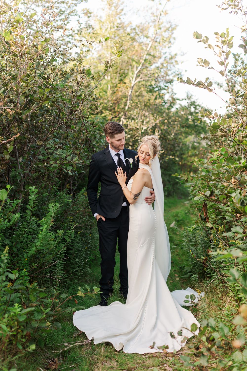 Bride and groom embrace in lush green garden, bride wearing elegant white gown with veil, groom in classic black suit.