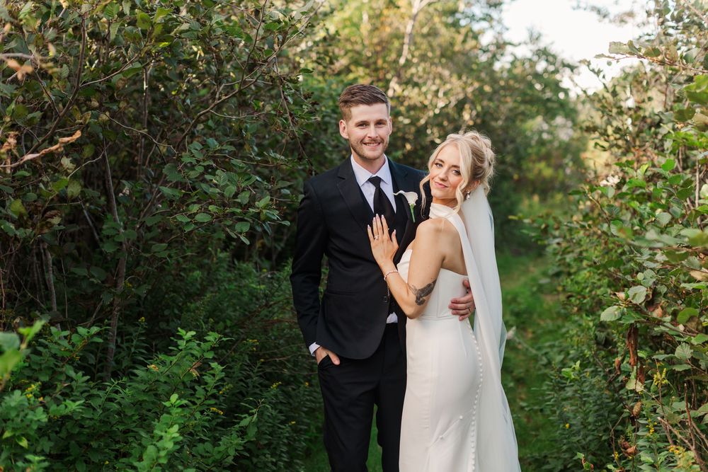 Bride and groom posing together in lush green garden, bride in white gown with veil, groom in black suit.