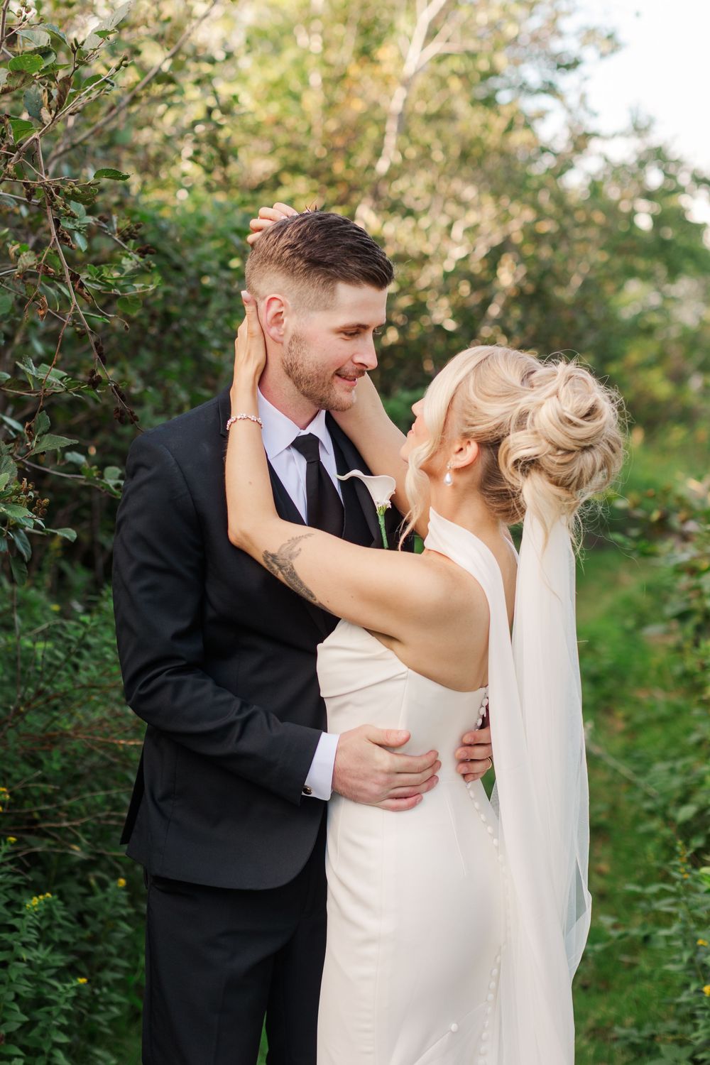A bride and groom share a romantic moment outdoors, surrounded by lush greenery, as she gently touches his face.