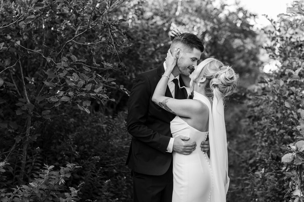 Black and white photo of a bride and groom sharing a romantic kiss surrounded by lush greenery on their wedding day.
