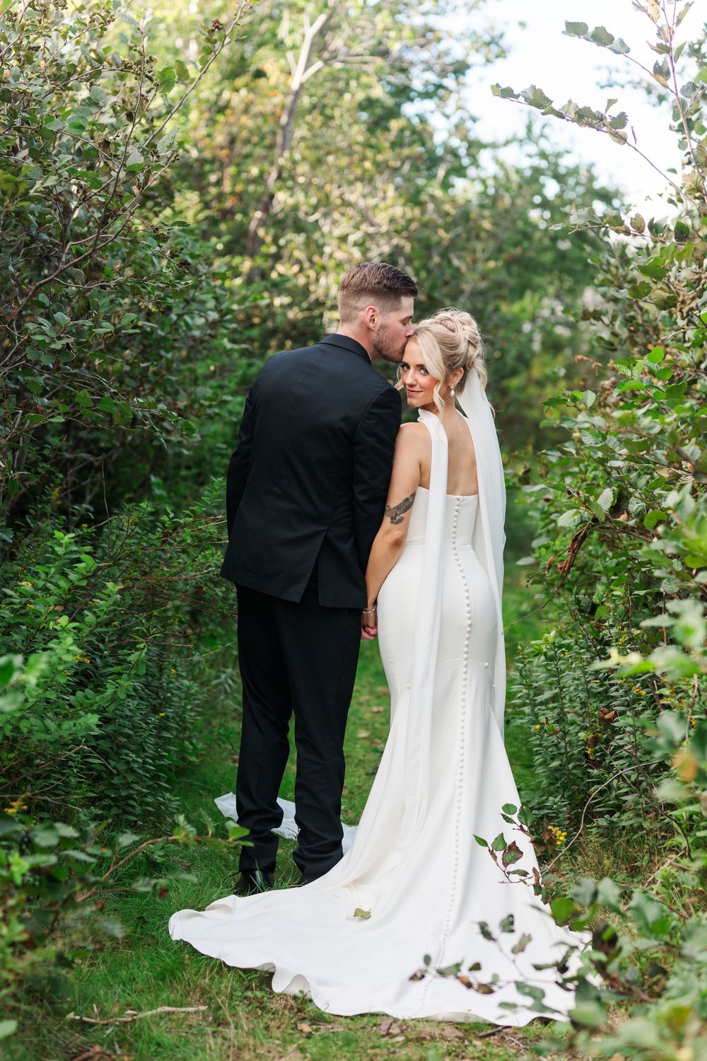 A bride and groom share a romantic moment in a lush green garden, bride wearing a white gown with button-back detail.