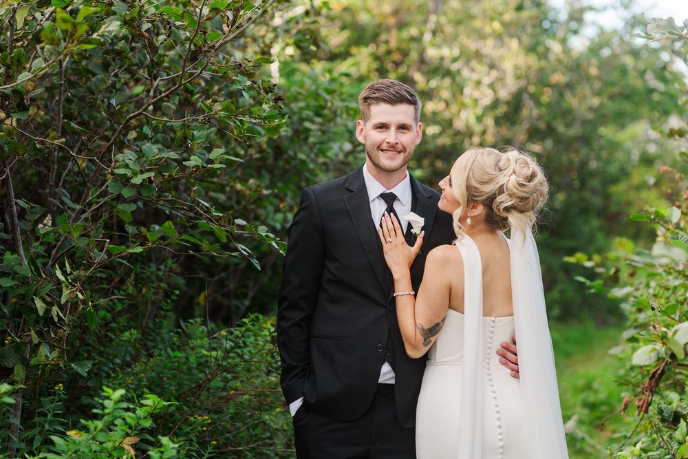 Bride and groom posing outdoors among lush green trees, smiling together in elegant wedding attire.