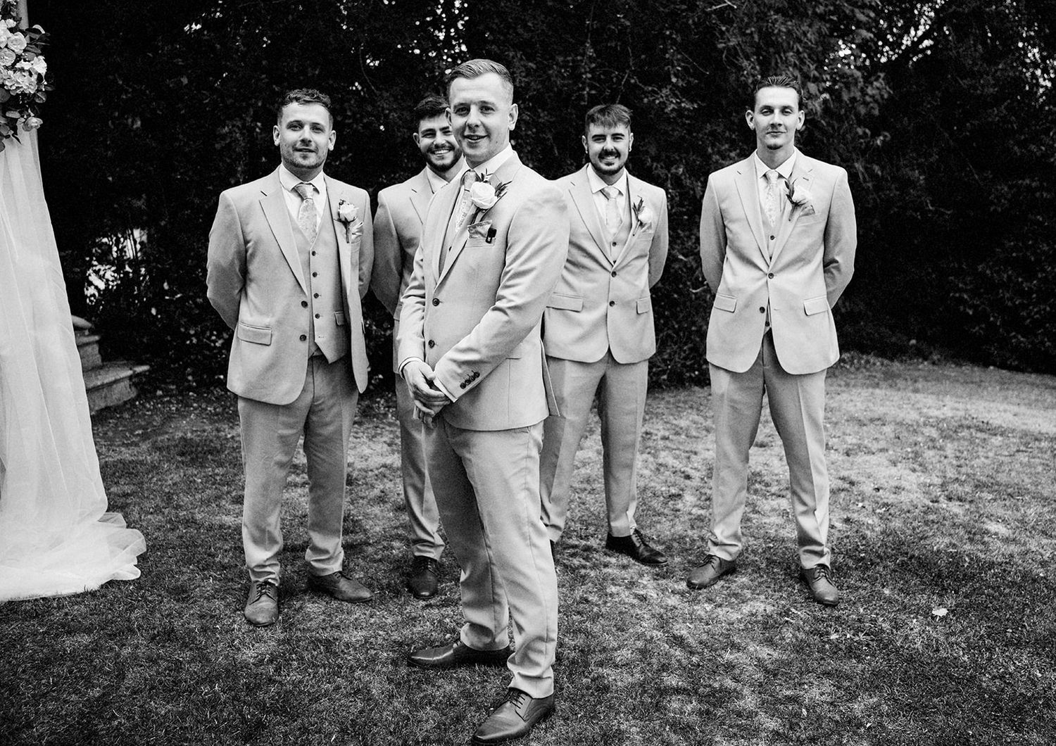Black and white photo of a groom and four groomsmen in matching suits posing outdoors at a wedding.