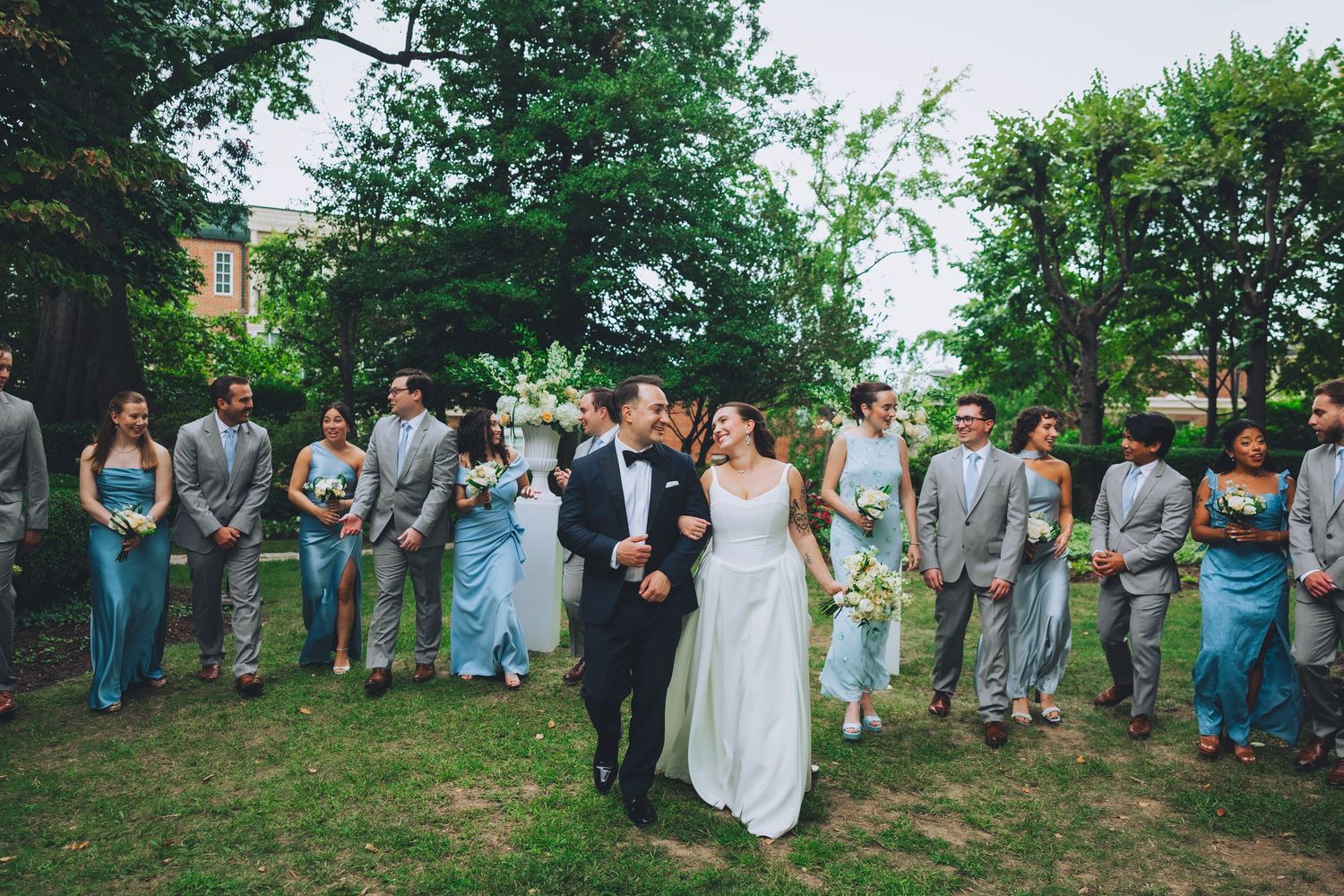 Bride and groom walk together outdoors surrounded by wedding party in blue dresses and gray suits on green lawn of the Meridian house in dc