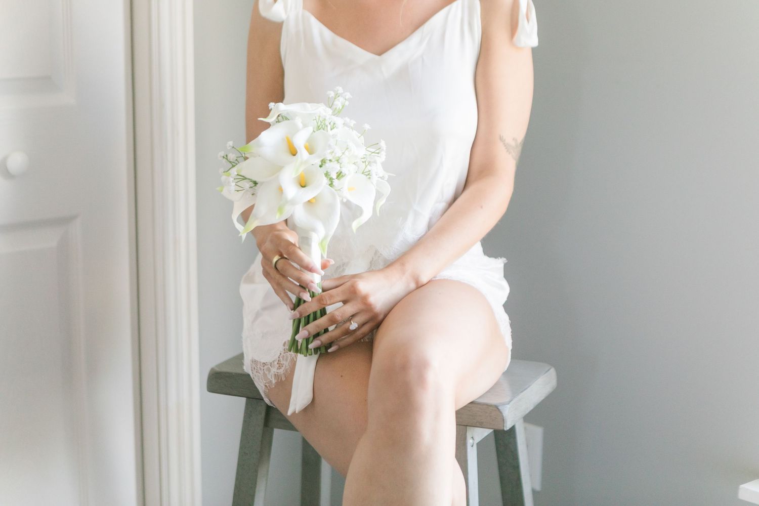 Bride in white dress sitting on stool holding elegant white calla lily bouquet, wedding preparation photo.