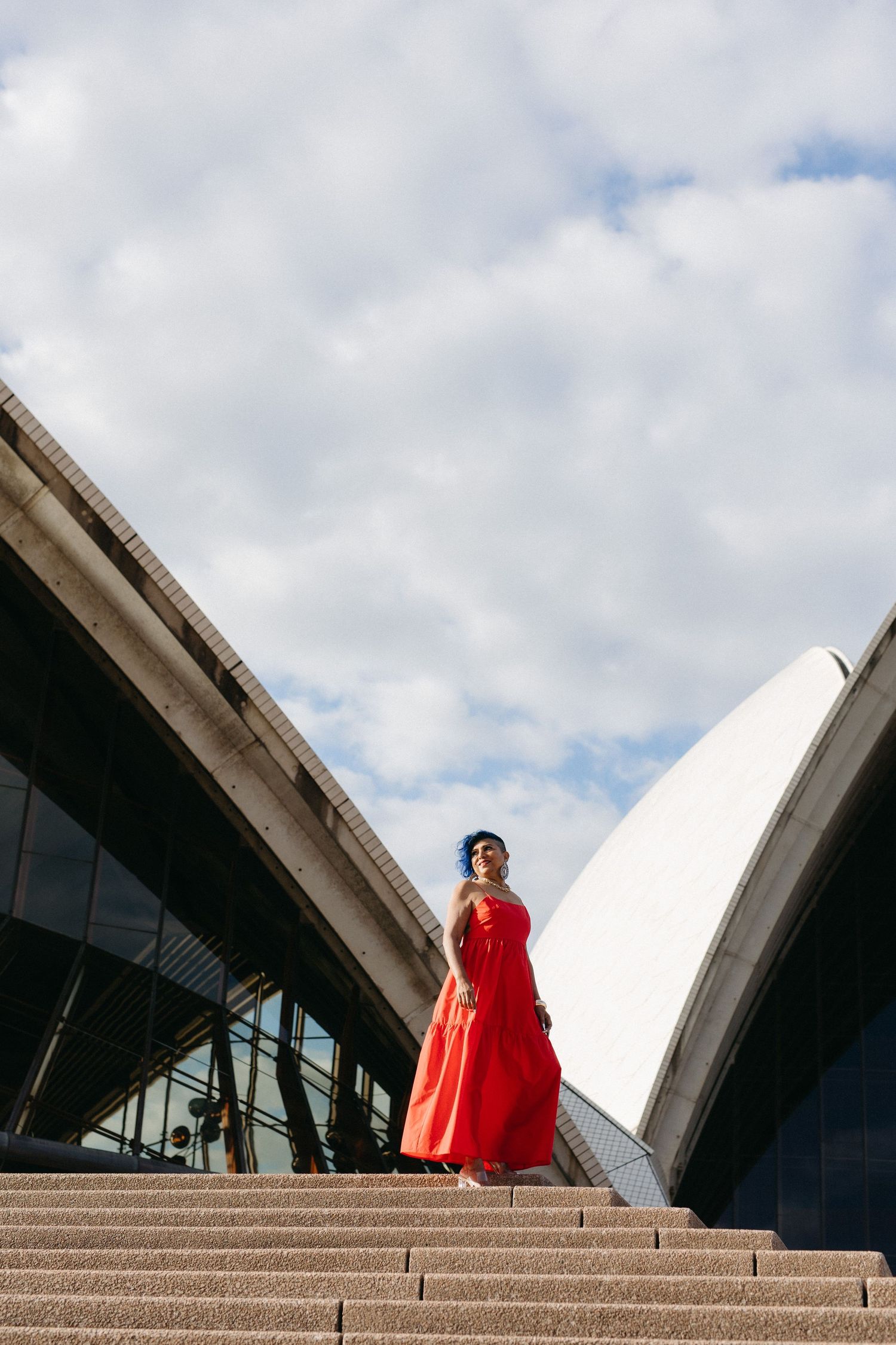 Woman in red gown poses on Sydney Opera House steps under cloudy blue sky.