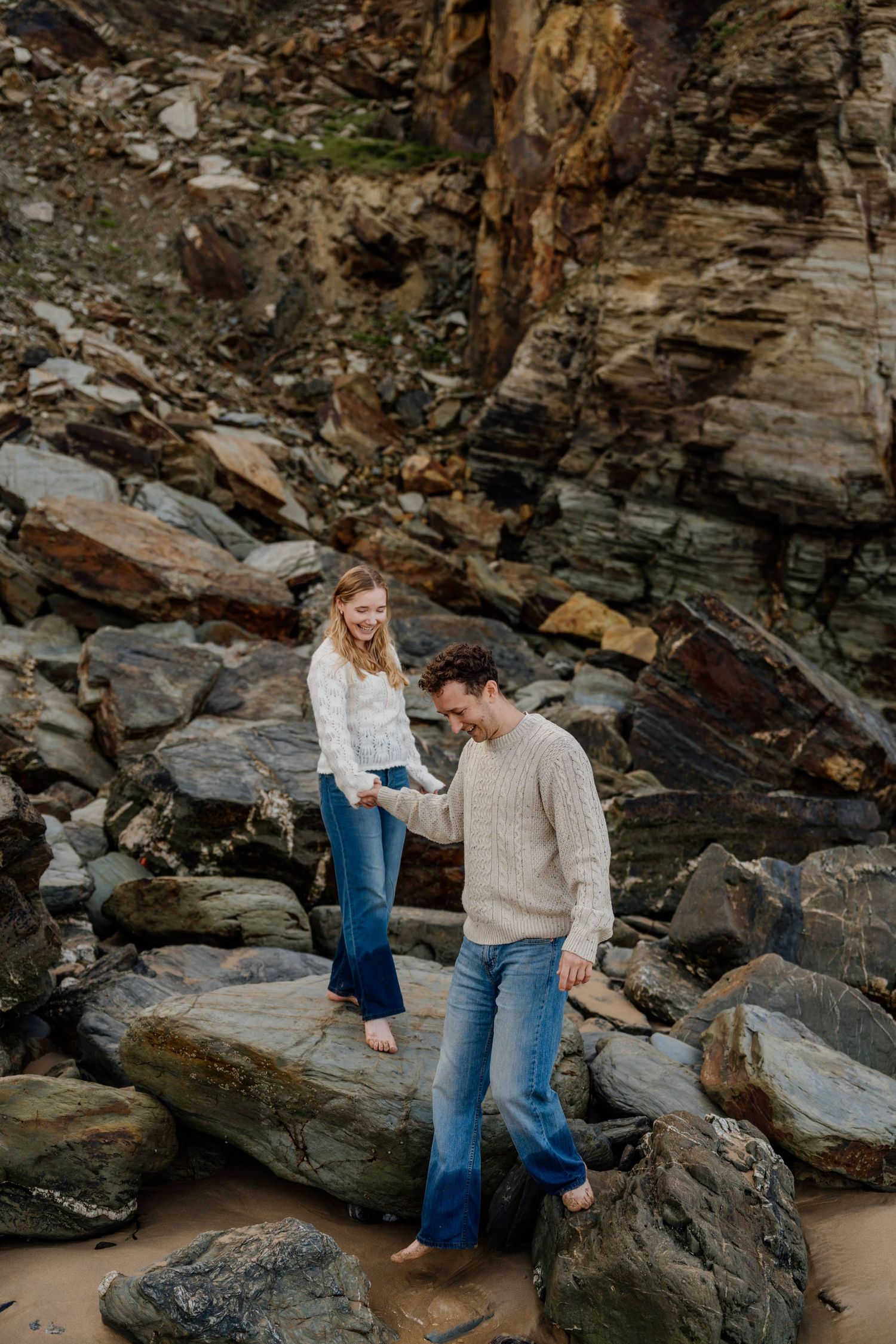 A couple holds hands while climbing rocks at watergate bay, wearing casual sweaters and jeans with rocky cliffs behind them.