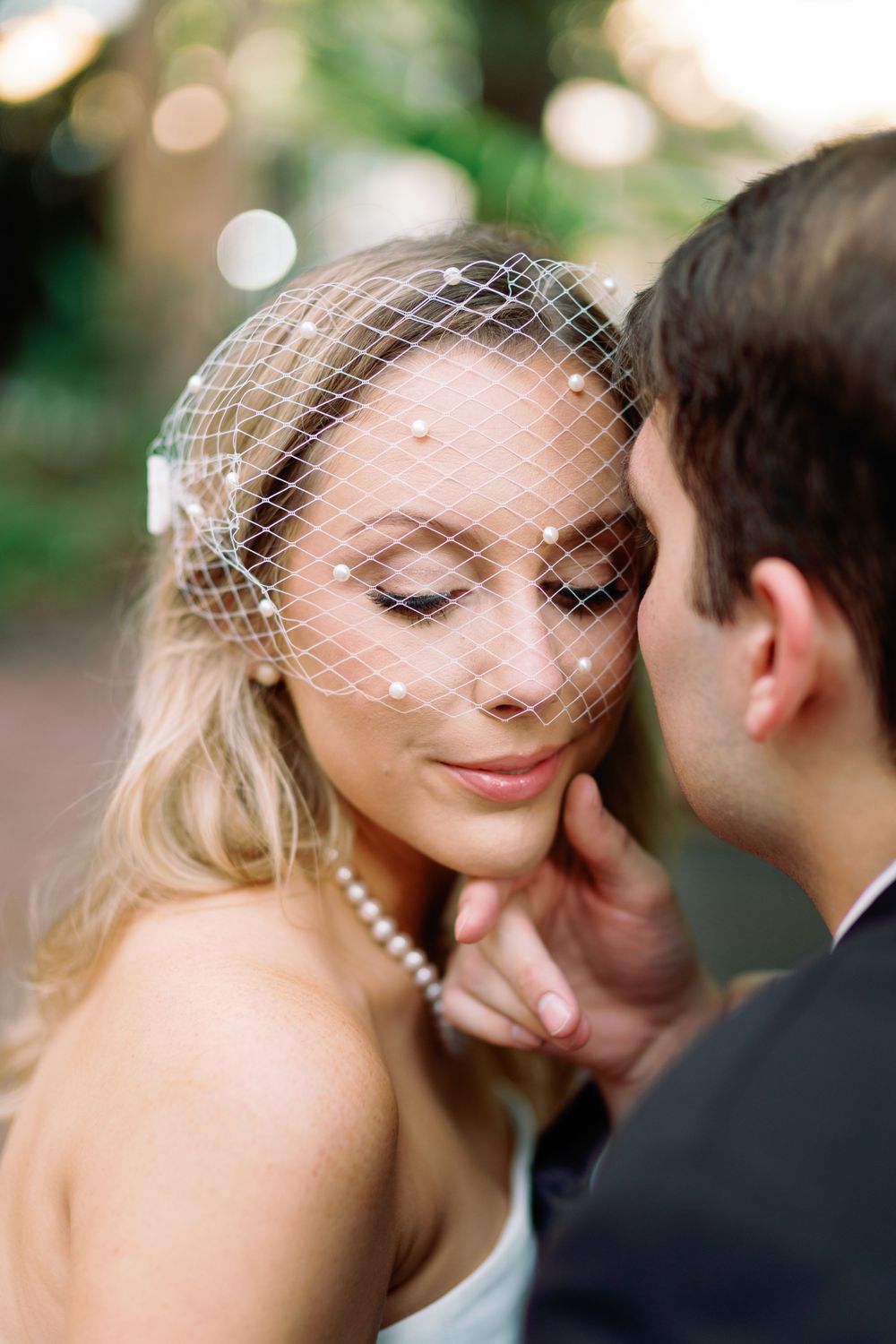 Close-up of blonde bride in pearl birdcage veil with eyes closed as groom tenderly touches her face.