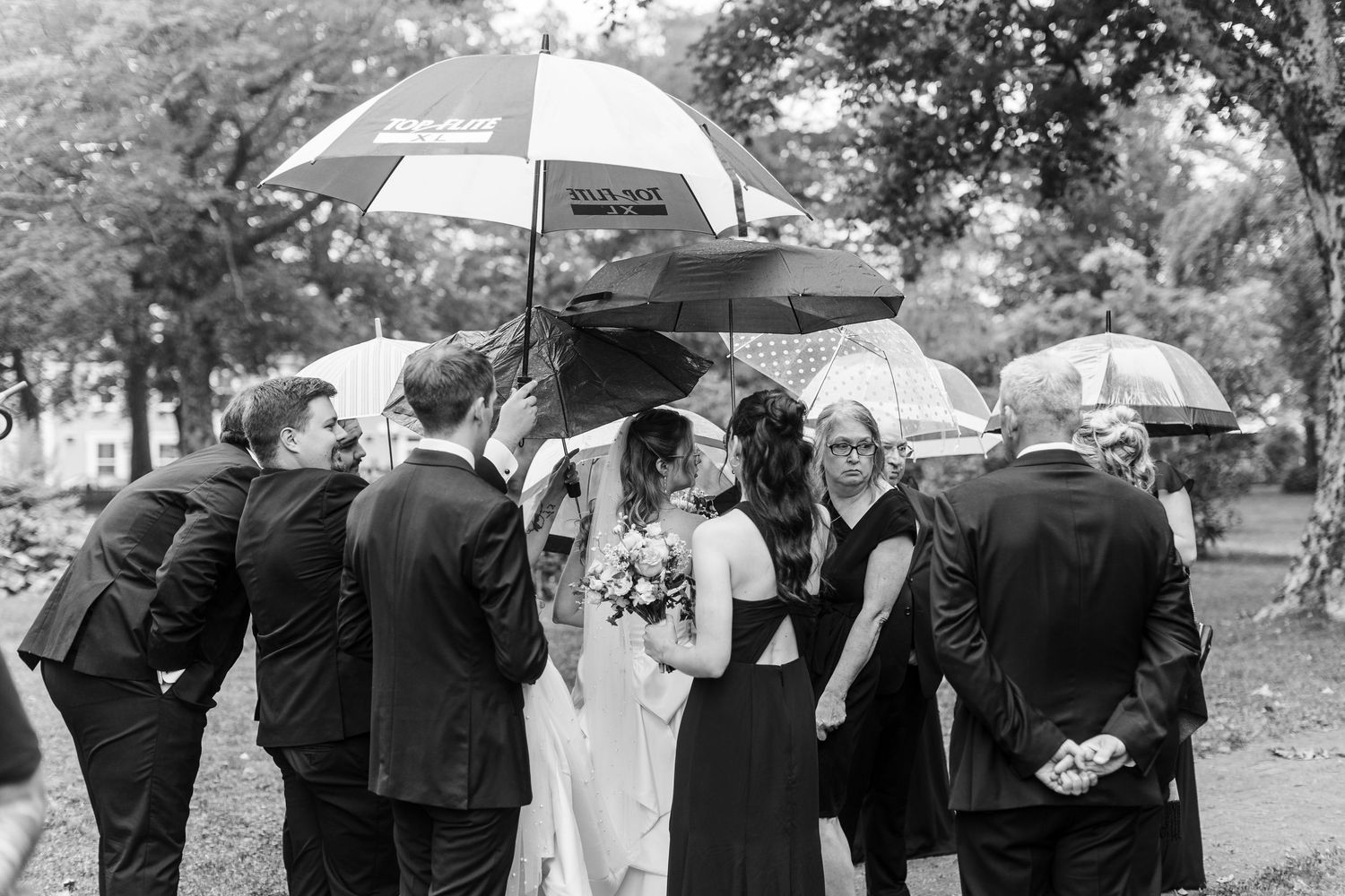 Black and white wedding photo of bride and groom kissing under umbrellas in the rain surrounded by guests.