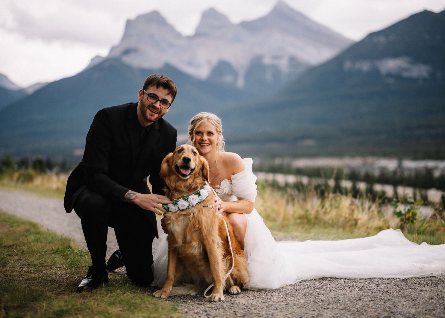 Bride and groom with their golden retriever in a white floral collar, Three Sisters behind, Canmore wedding