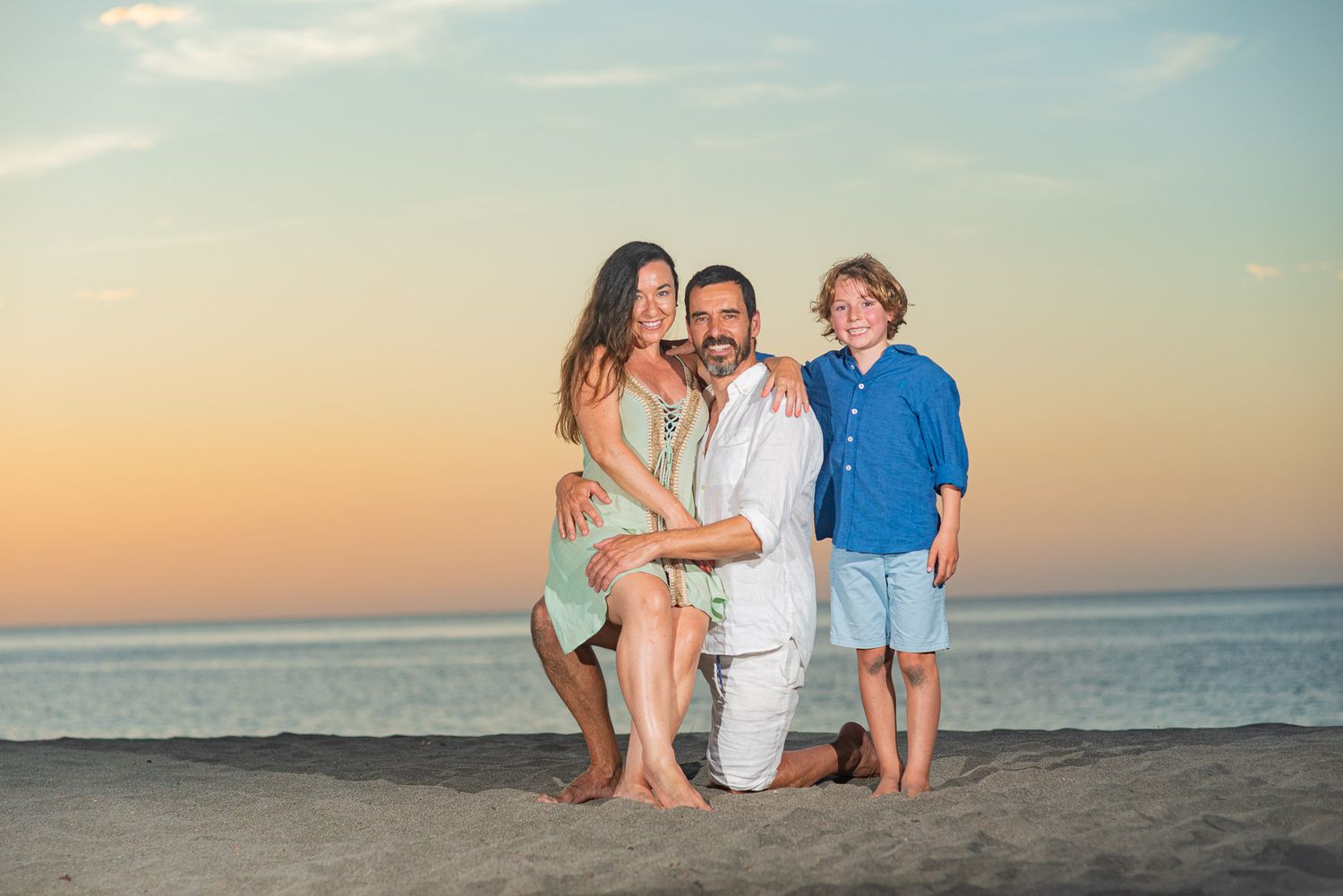 Family of three posing on sandy beach at sunset, parents and young boy smiling in coordinated outfits.