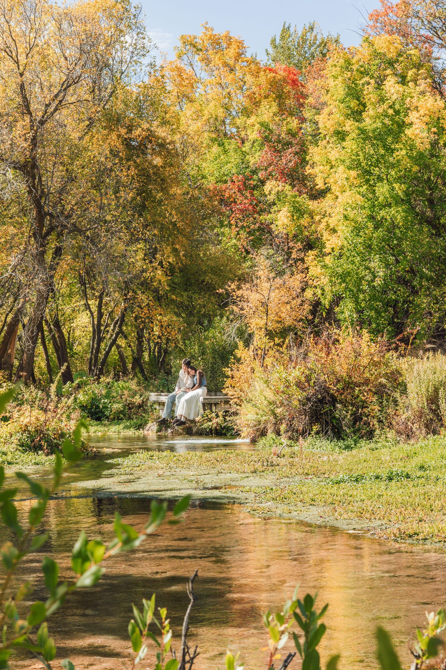 Couple sitting on a dock by a tranquil pond surrounded by vibrant autumn foliage in golden and red hues.