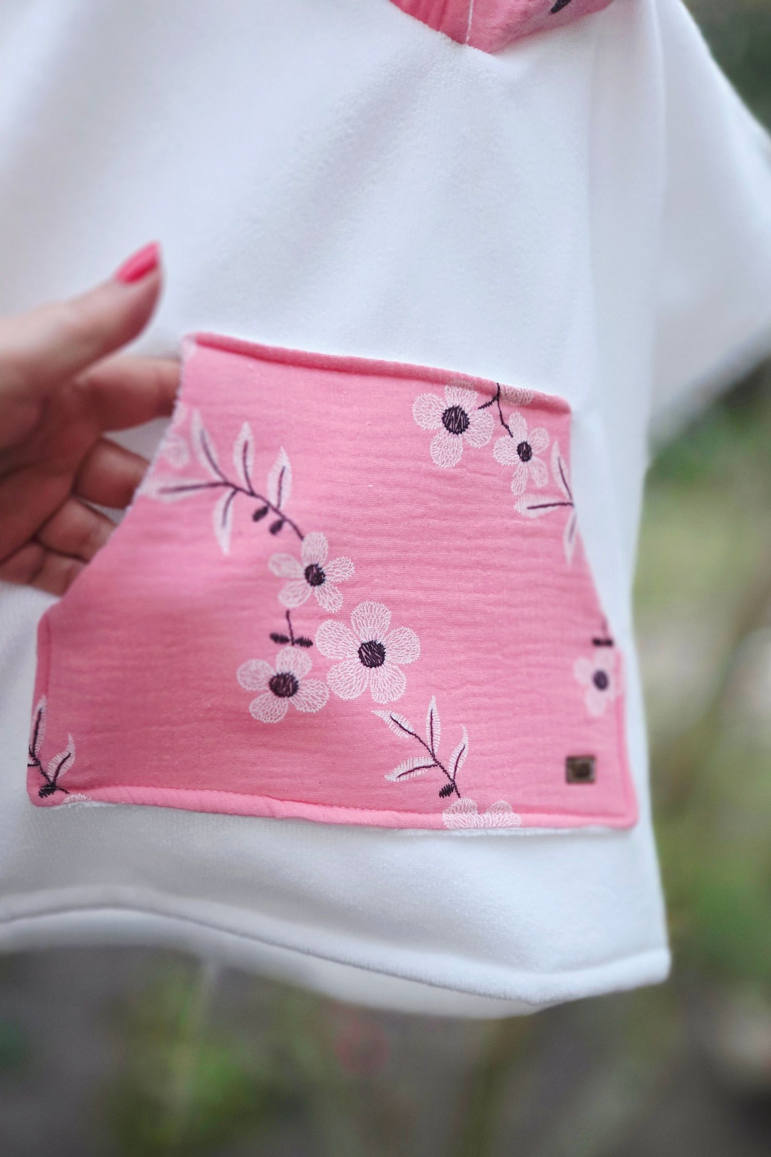 Close-up of a white hoodie with a pink floral pocket featuring cherry blossom embroidery, held by a hand with pink nails.