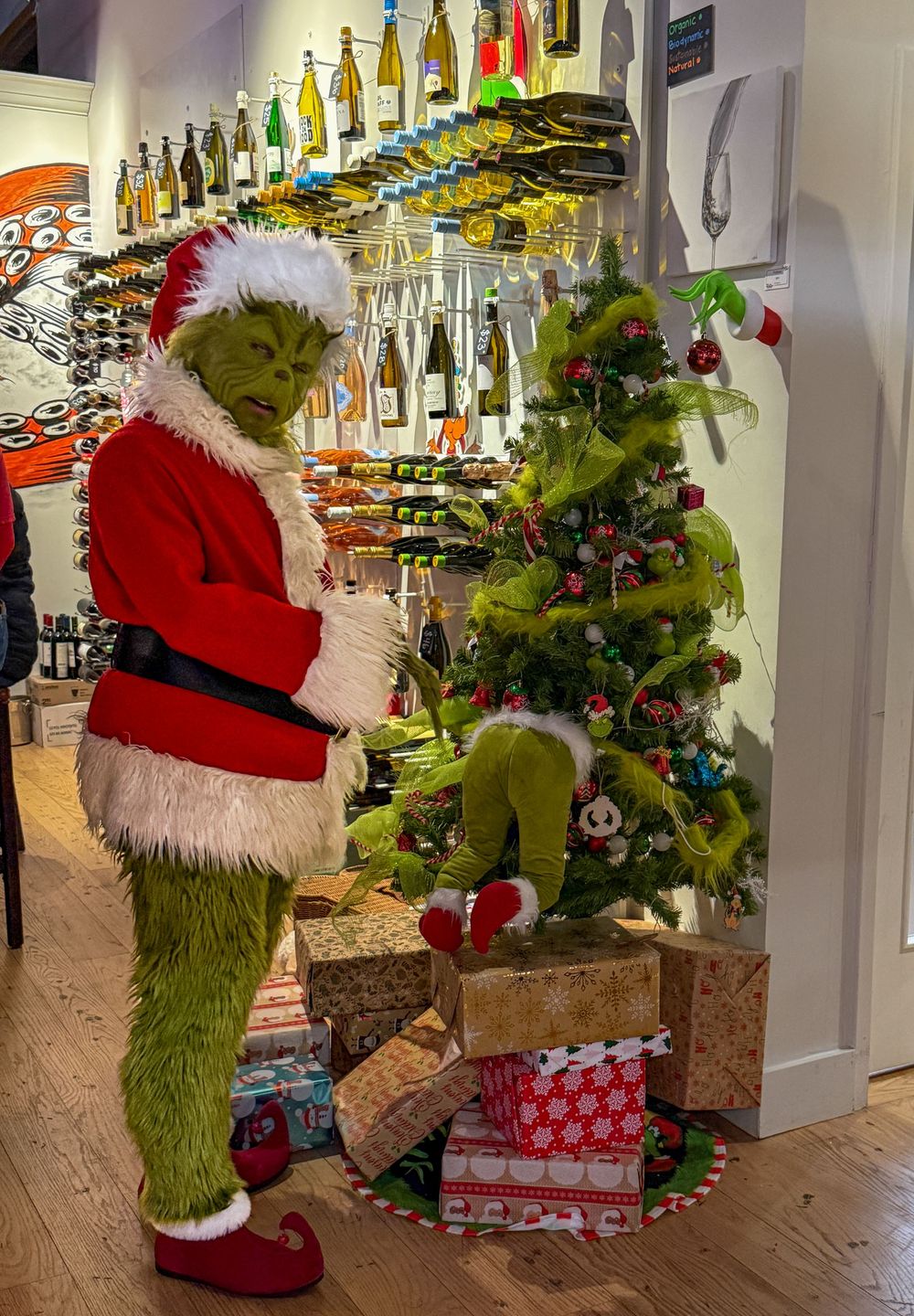 Grinch character in Santa suit standing beside decorated Christmas tree with wrapped gifts in wine shop.