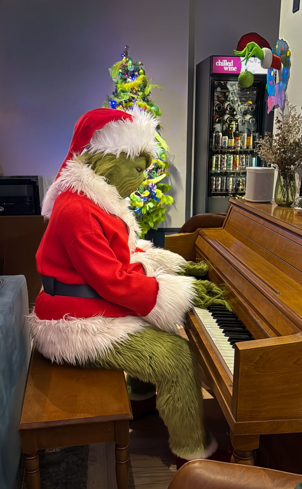 The Grinch dressed in Santa costume playing upright piano beside a decorated Christmas tree.