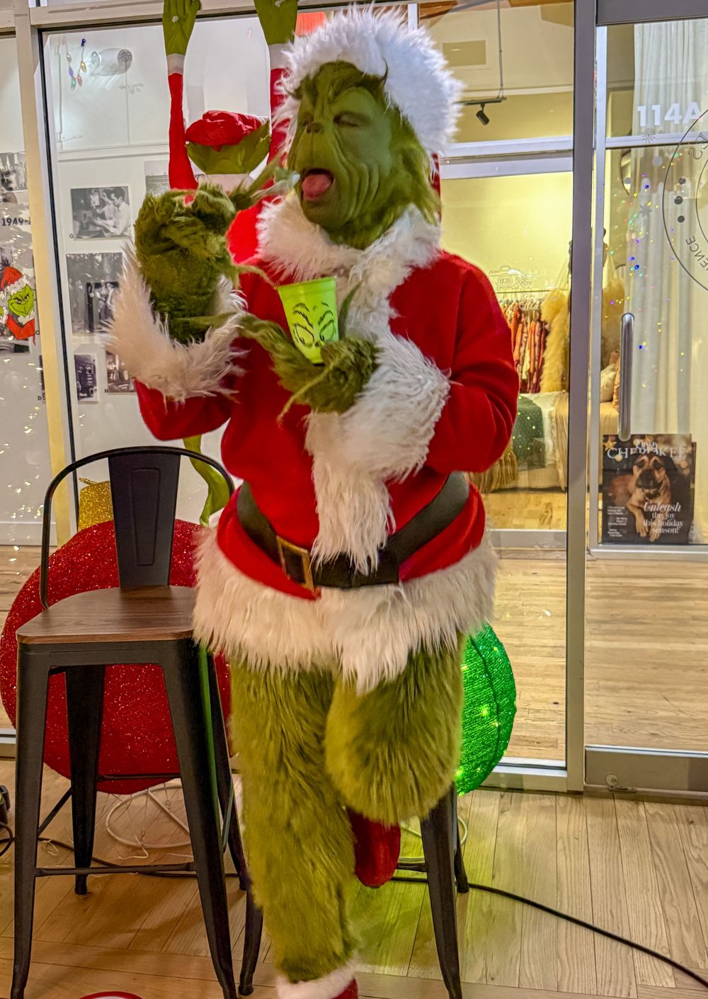 Grinch in Santa outfit holding green mug with tongue out, posing playfully in festive decorated store.