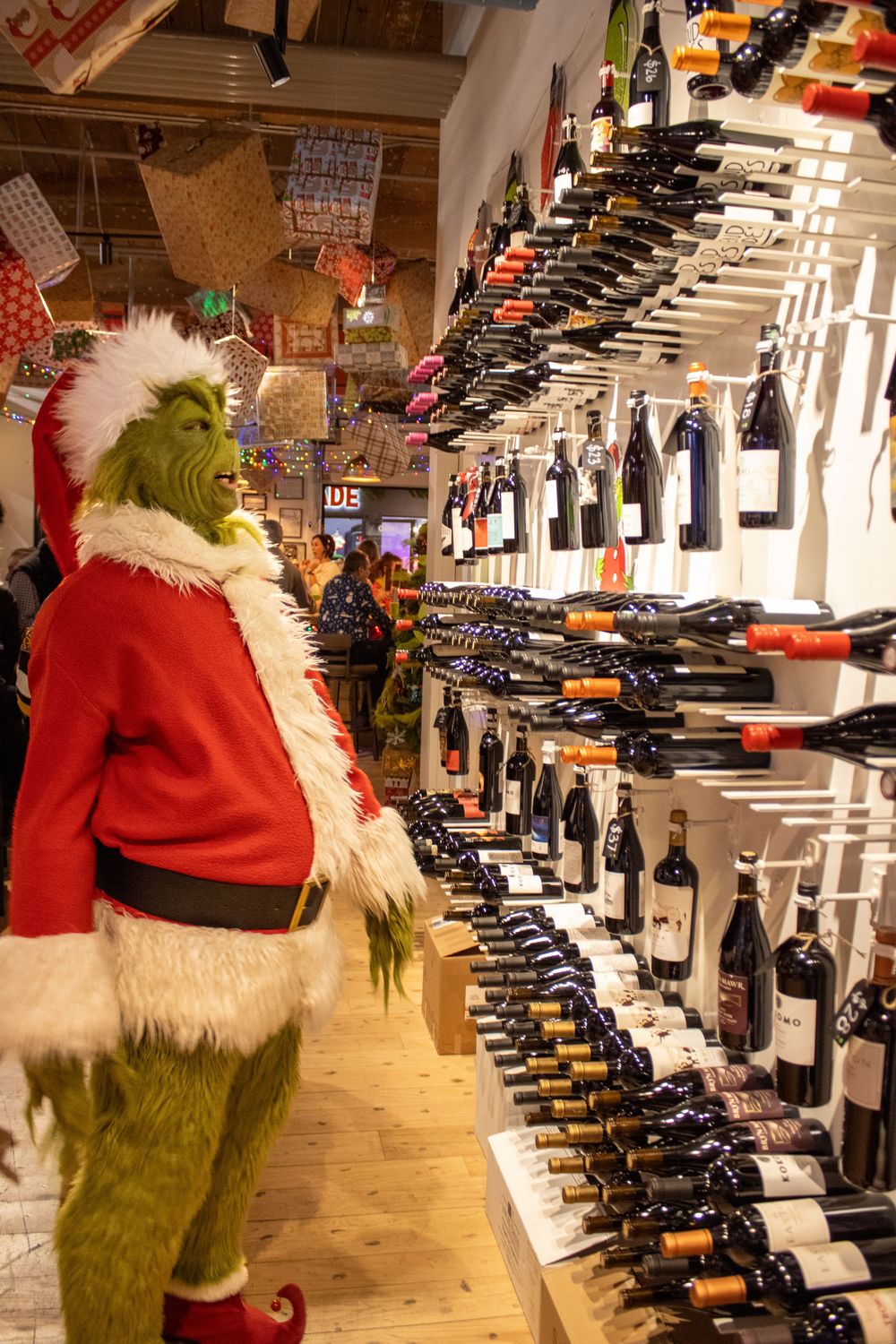 Grinch character in Santa costume browsing wine bottles on display shelves in holiday decorated shop.