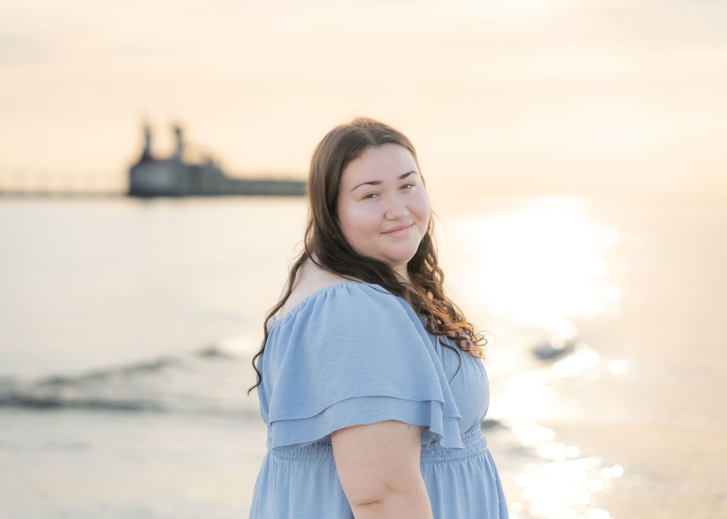 Young woman in blue dress smiling at beach during golden sunset with ship silhouette in background.
