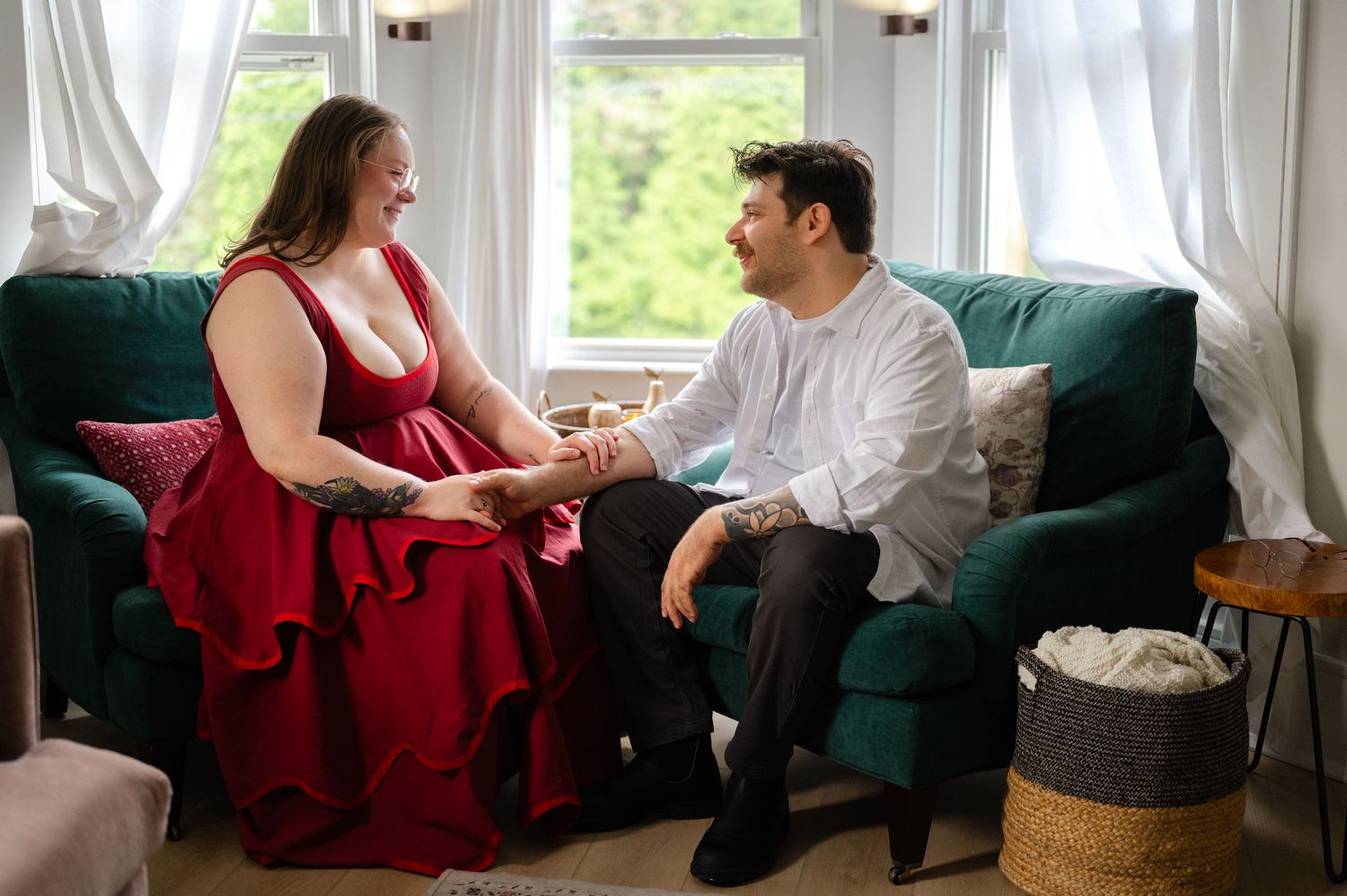 Couple in formal attire holding hands on a green sofa, woman in red dress, man in white shirt. NJ home during an engagement session.
