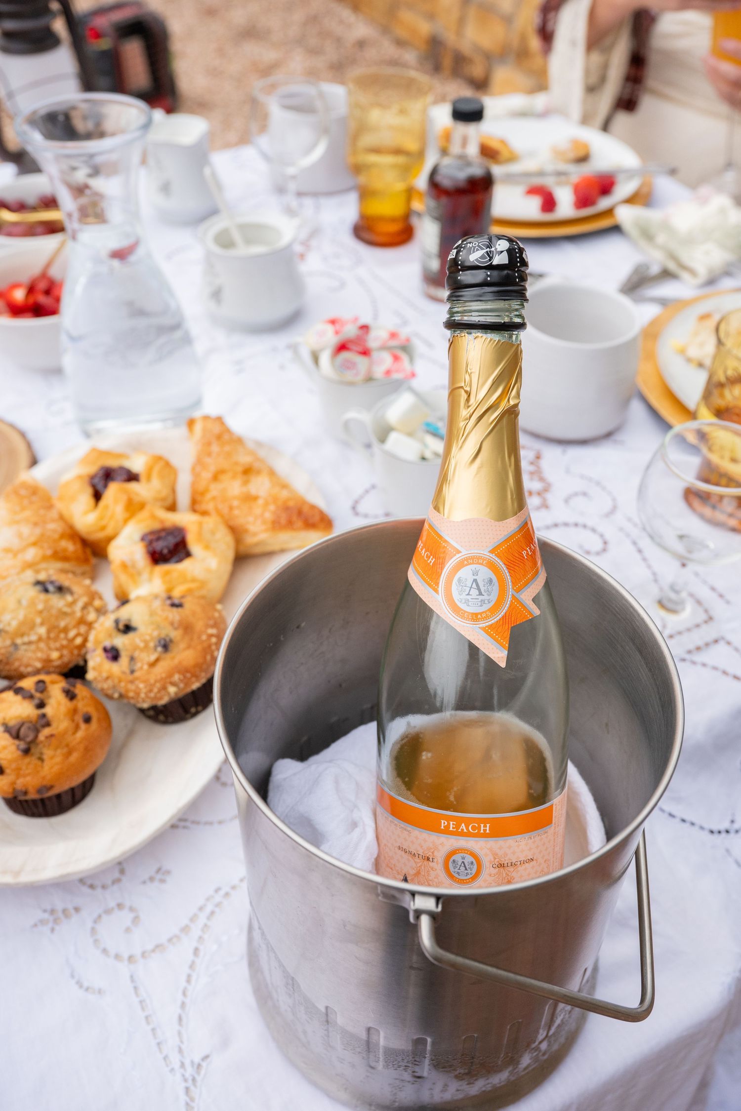 Peach sparkling wine chilling in ice bucket at brunch table with muffins and pastries.