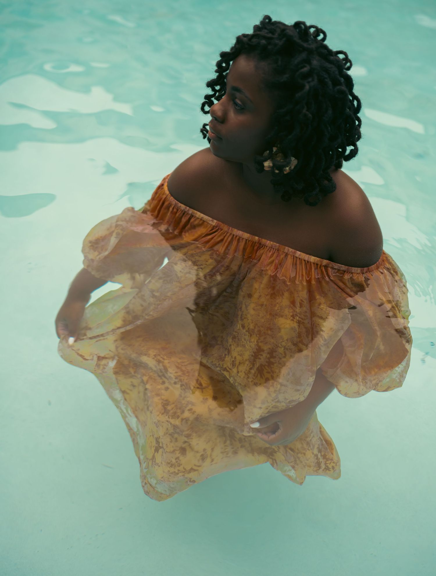 Woman in orange off-shoulder dress standing in turquoise pool water, fabric flowing around her.