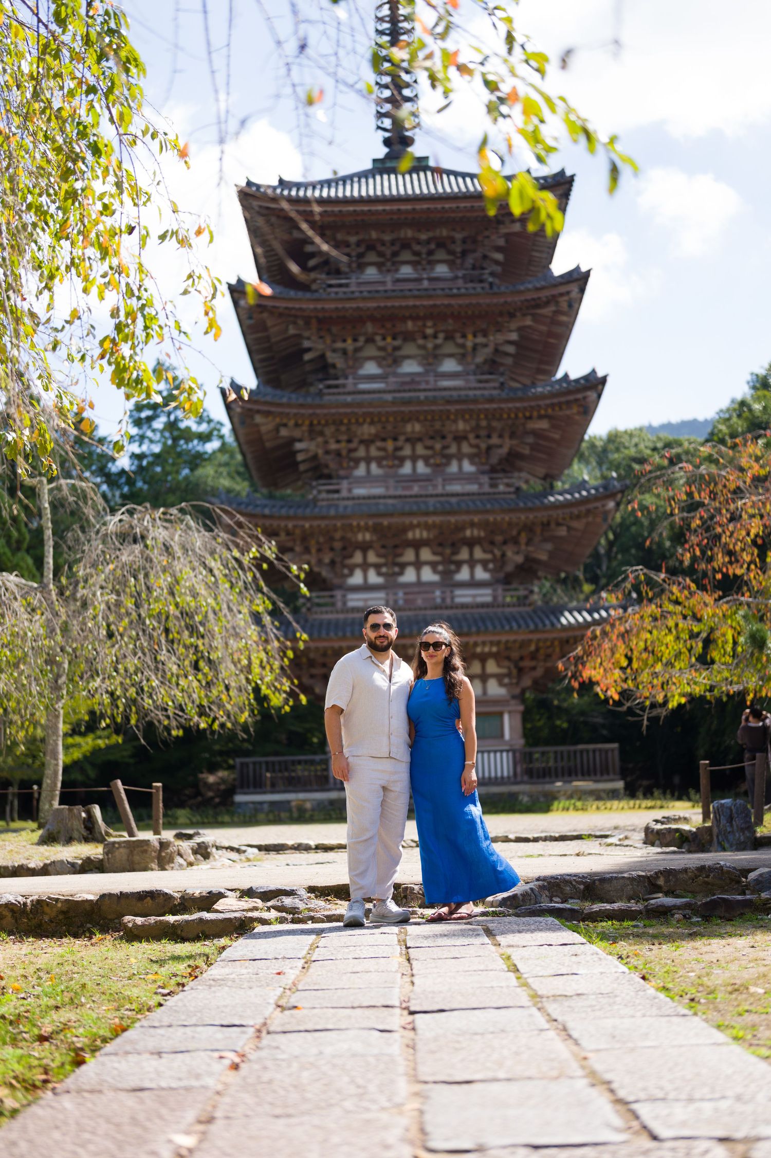 Couple in sunglasses posing on a stone path in front of a historic multi-tiered Japanese pagoda surrounded by autumn trees.