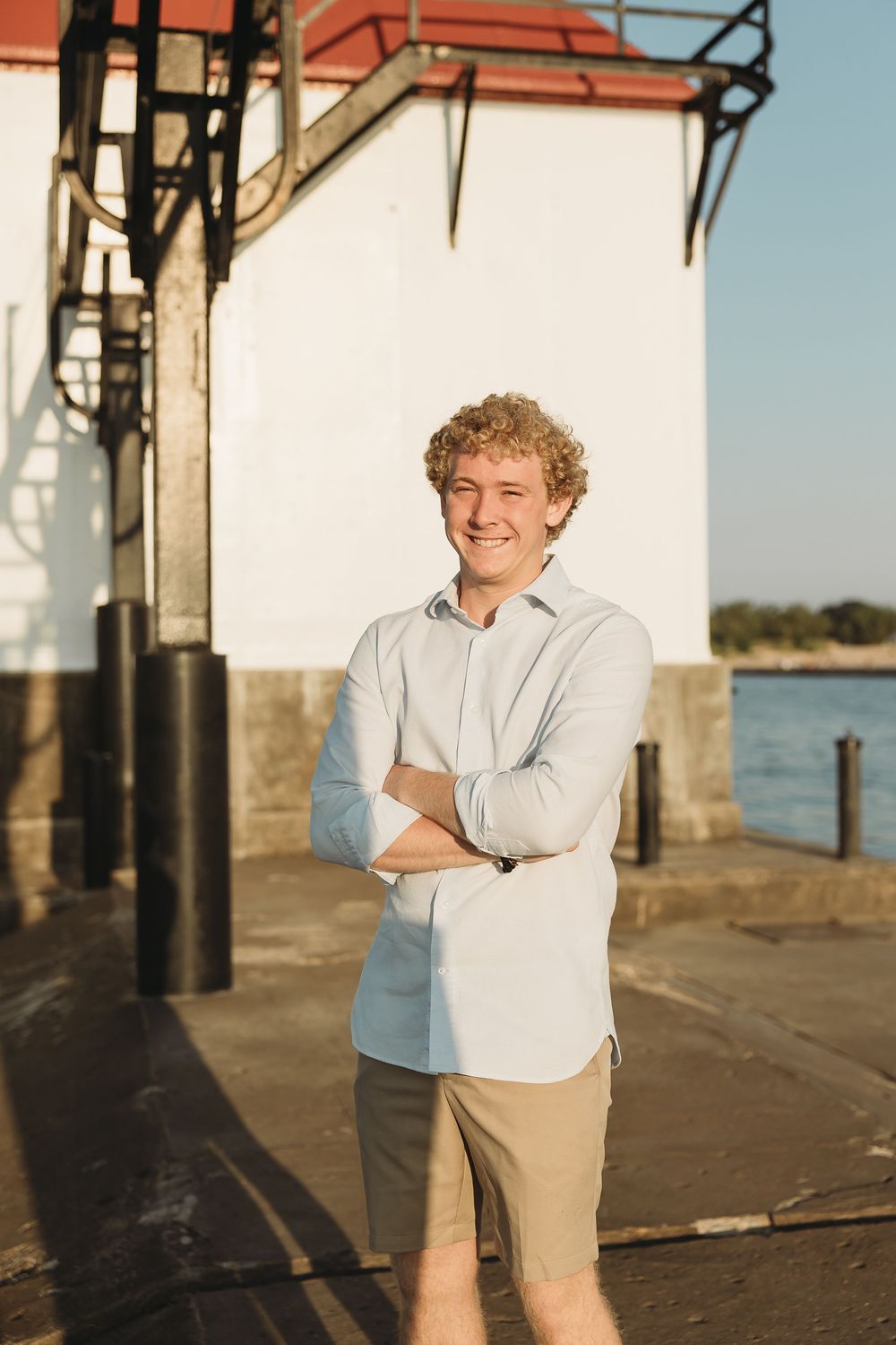Smiling young man with curly hair stands with arms crossed in a light blue shirt and khaki shorts beside a white lighthouse at golden hour.