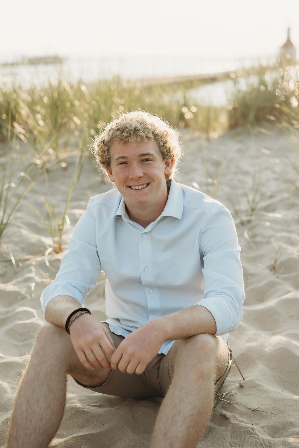 Smiling young man with curly hair in a light blue shirt sits on a sandy beach with dune grass and a calm shoreline behind him.