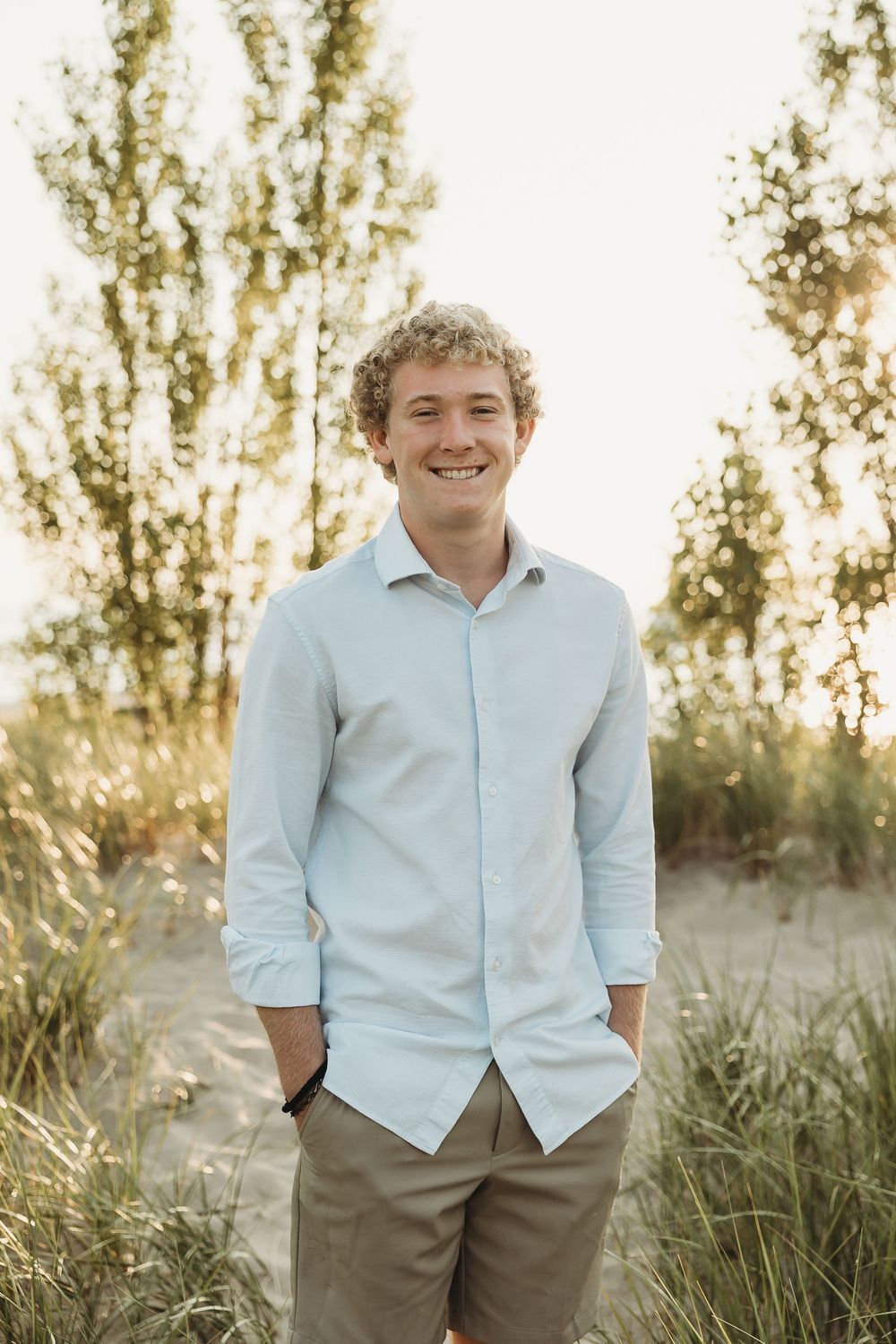 Smiling young man with curly hair in a light blue button-up shirt and khaki shorts standing on a sandy path lined with tall grass and trees.