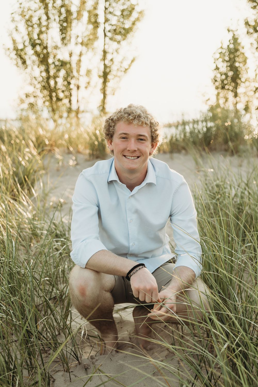 Smiling young man with curly hair in a light blue button-up shirt crouches among tall beach grass on a sandy dune at sunset.