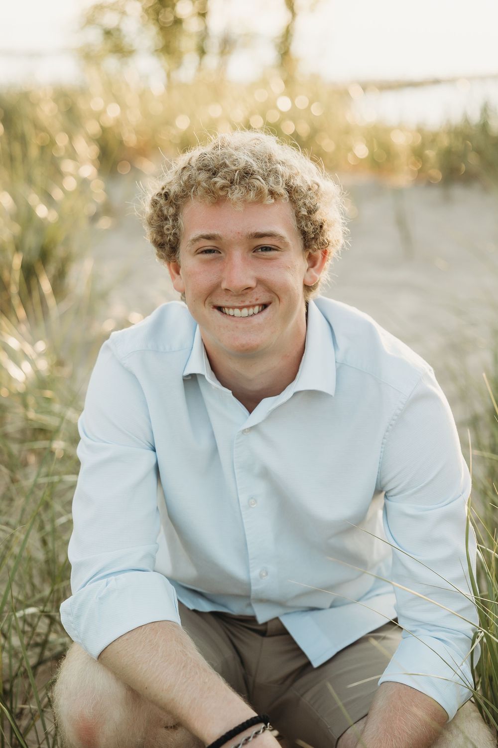 Smiling young man with curly hair in a light blue button-up shirt and khaki shorts crouching among beach grass at sunset.