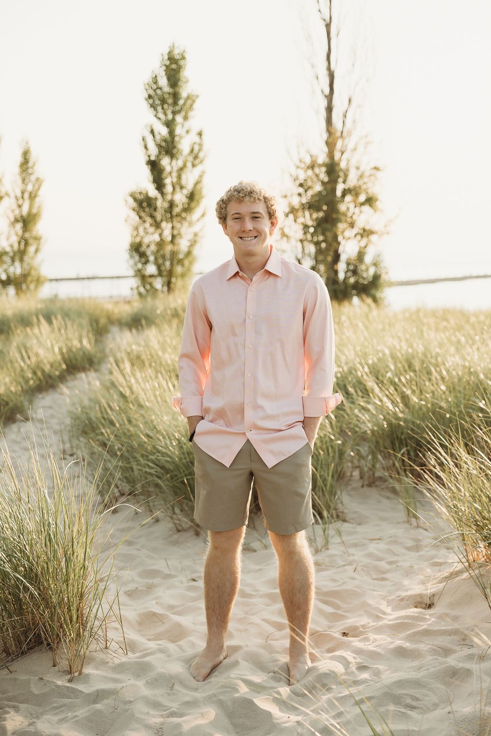 Smiling young man in a pink button-up shirt and khaki shorts stands barefoot on a sandy beach path lined with tall dune grass at sunset.