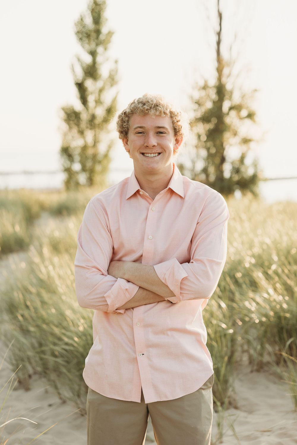 Smiling young man with curly hair and arms crossed, wearing a pink button-up shirt, standing in beach grass at sunset.
