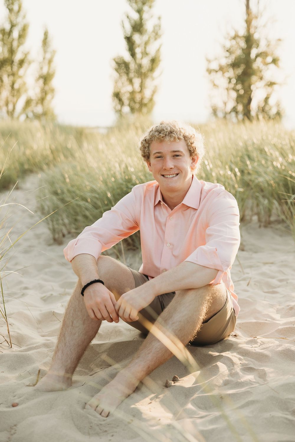 Smiling young man with curly blond hair in a coral button-up shirt and khaki shorts sitting barefoot on a sandy beach dune with tall grass.