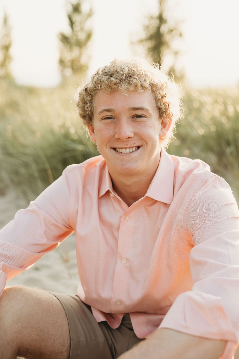 Smiling young man with curly blond hair in a coral button-up shirt sits on a grassy beach dune during golden hour.