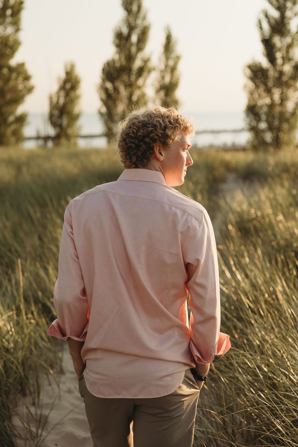 Curly-haired young man in a pink shirt stands with his back to the camera on a dune path lined with tall grass and evergreen trees.