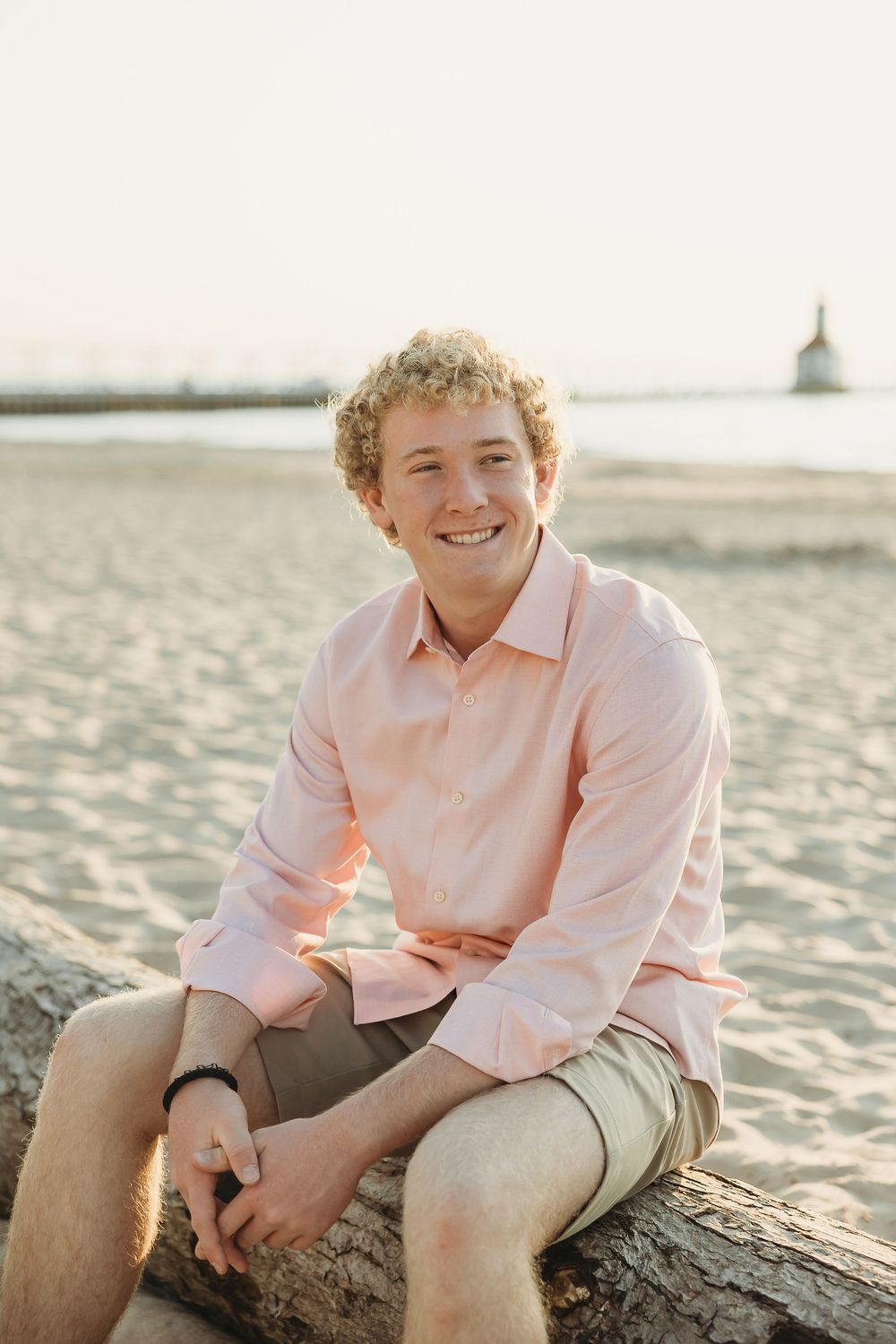Smiling young man with curly blond hair in a pink button-up shirt sits on a driftwood log on a sandy beach with a lighthouse behind.