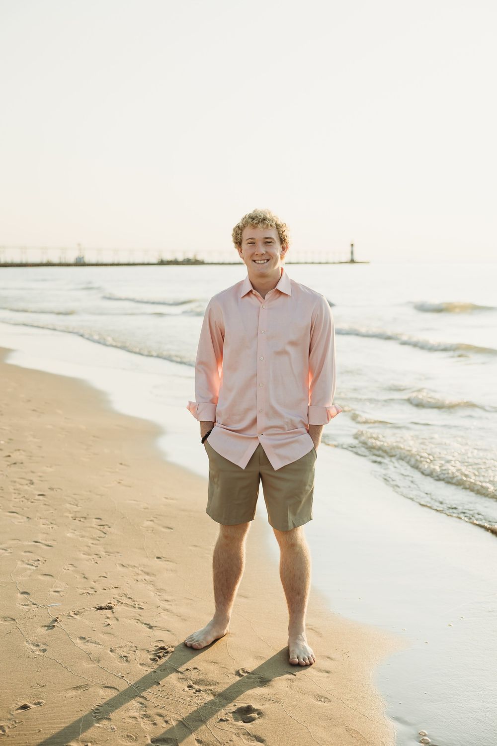 Smiling young man in a pink button-up shirt and khaki shorts stands barefoot on a sandy beach with a pier and lighthouse behind him.