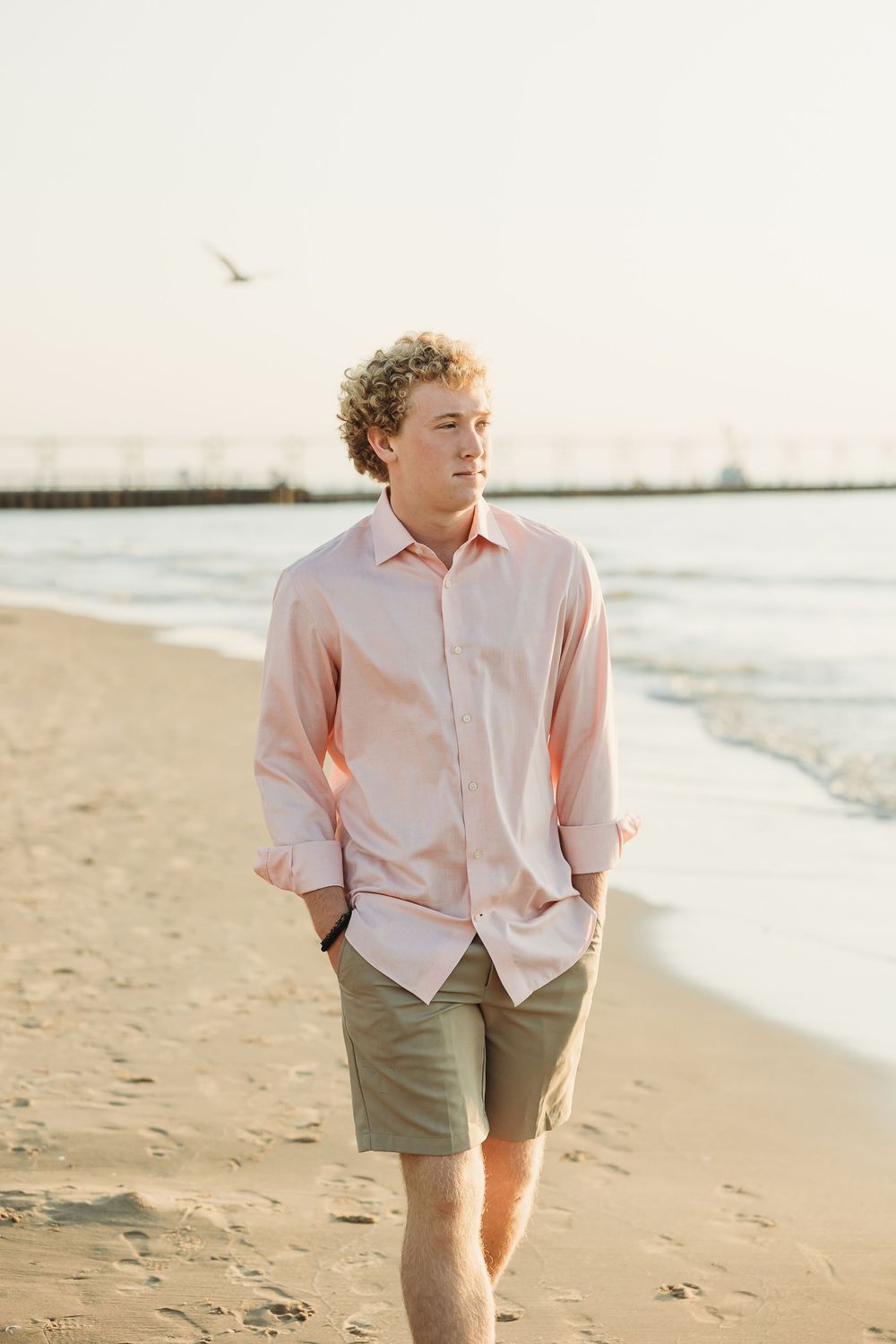 Young man with curly hair in a pink button-up shirt and khaki shorts walks along a sandy beach at sunset with a pier behind him.